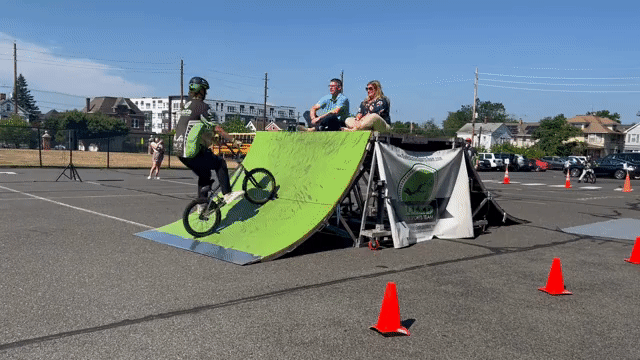 animated gif of a man and woman sitting on top of a bright green ramp as a kid on a bmx bike jumps over them